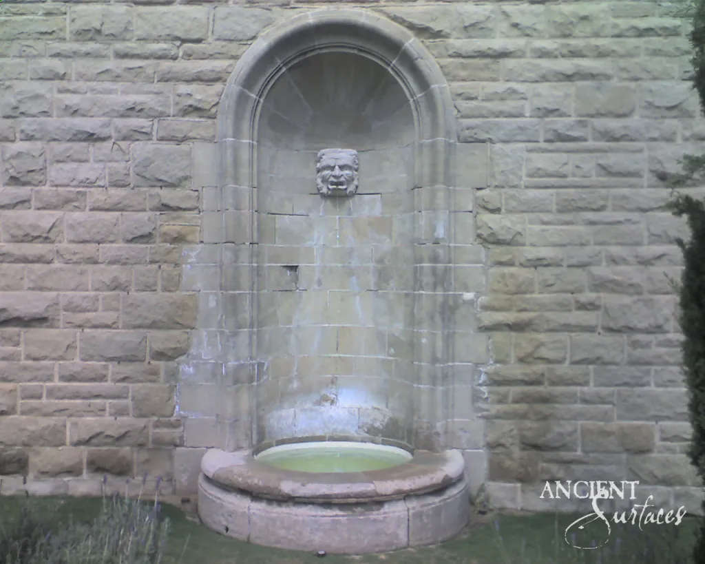 Arched stone wall fountain with a carved face mascaron sheeting water into a round basin